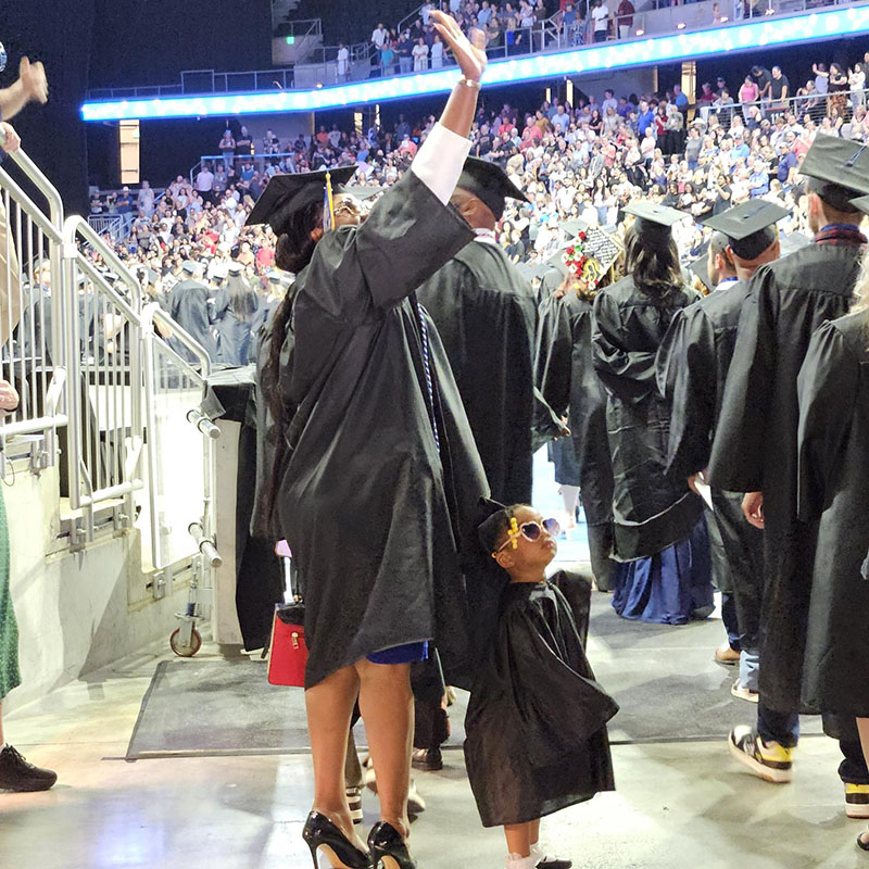 A mother celebrates graduation with her daughter, both dressed in gap and gown, waving to seated guests from the arena floor.